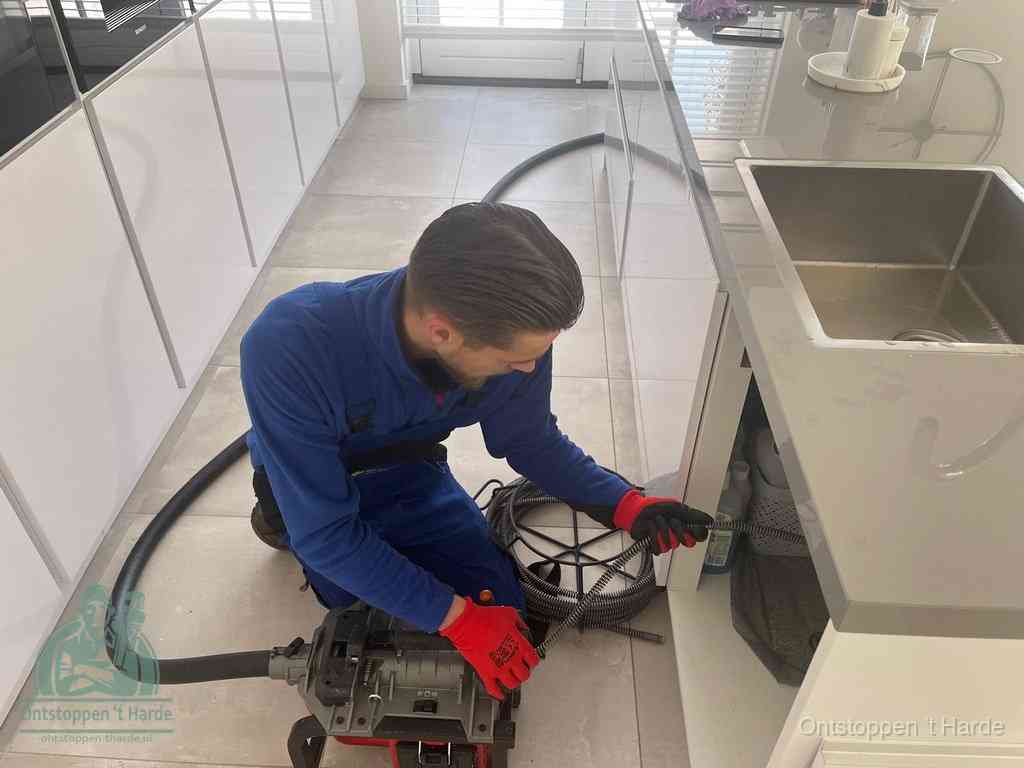 Een monteur ontstopt de keukenafvoer onder de spoelbak met een elektrische veermachine in t Harde