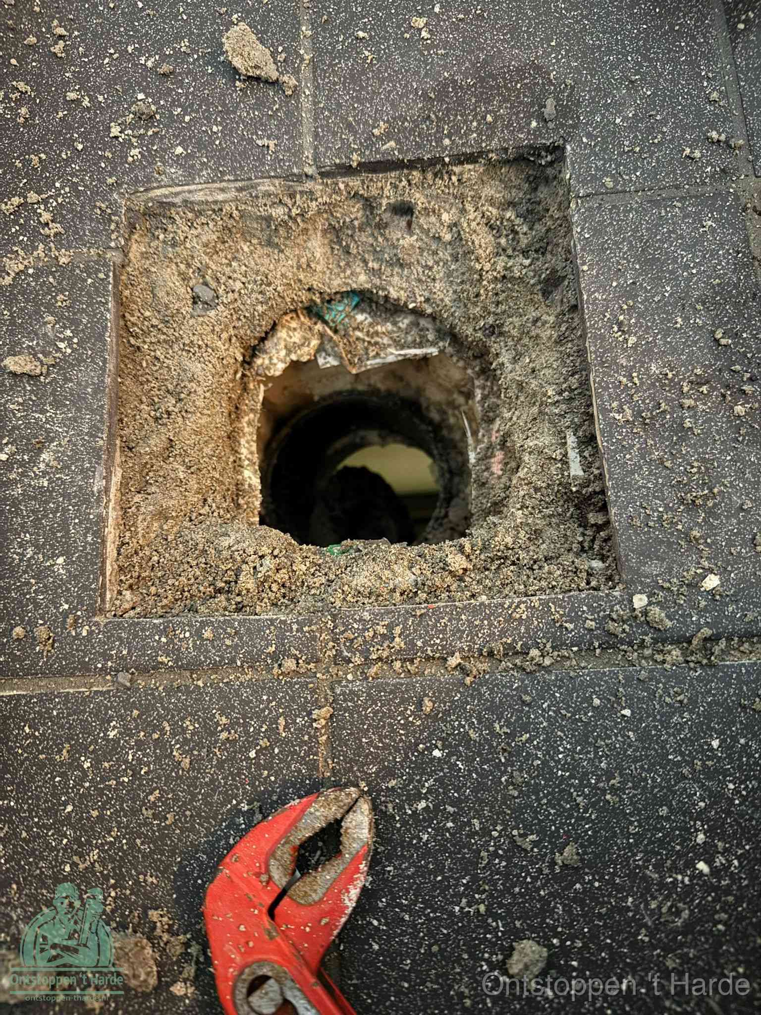Een geopend rioolputje in een betontegelvloer met losgemaakt rooster en een zichtbare ronde afvoerbuis, het achterhalen waar de afvoerverstopping zit in t Harde
