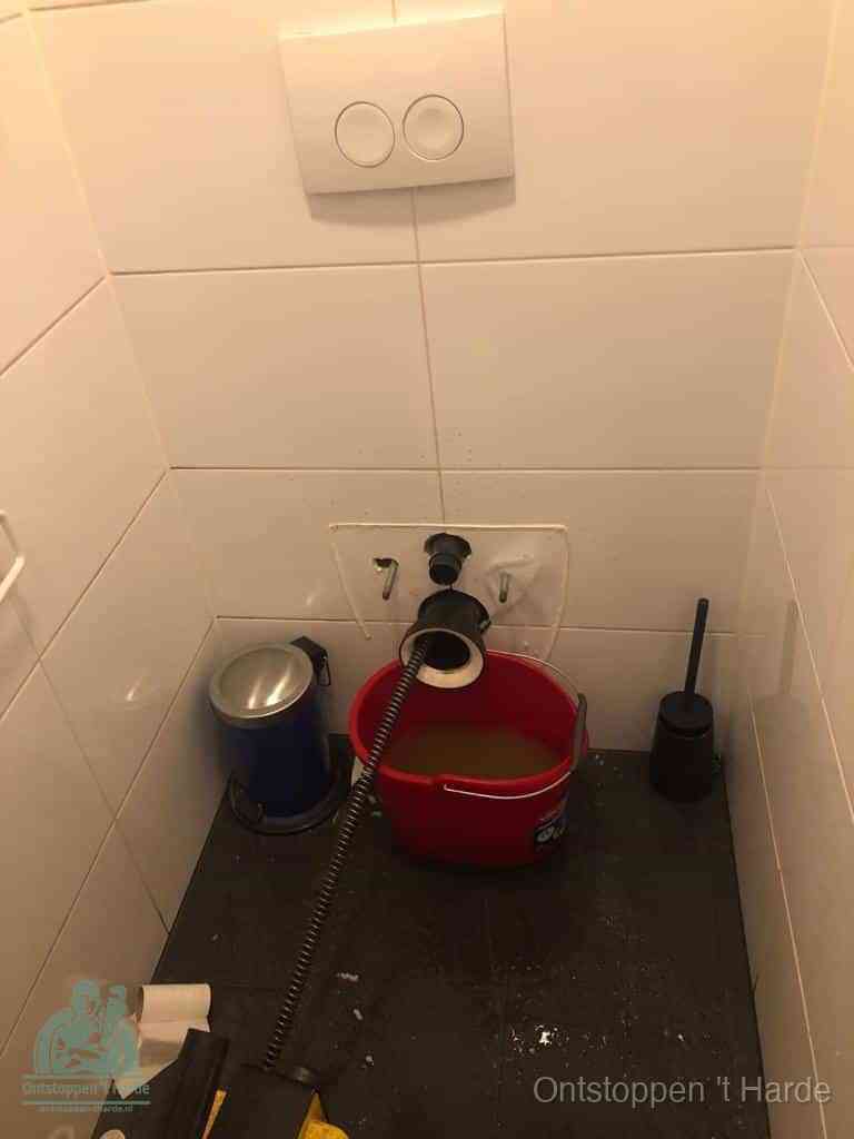 Een ontstopte wand-wc met verwijderde toiletpot waar een ontstoppingsveer in de wanduitgang verdwijnt, het achterhalen waar de verstopping zit in t Harde