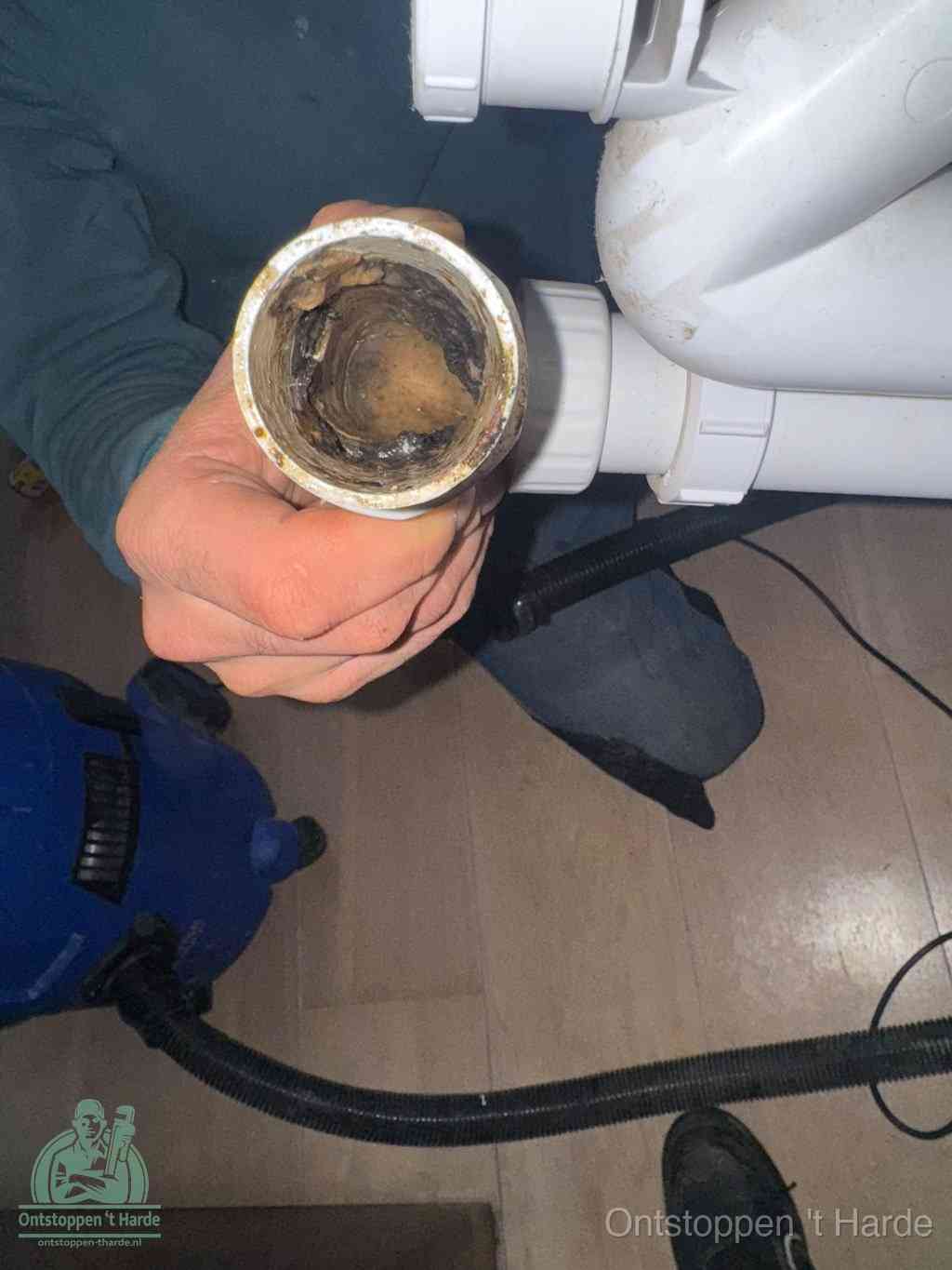 Een hand houdt een losgekoppeld PVC-afvoerbuisstuk vast dat van binnen volledig is dichtgeslibd met vet, vuil en aanslag, de meest voorkomende oorzaak van een verstopte afvoer in t Harde