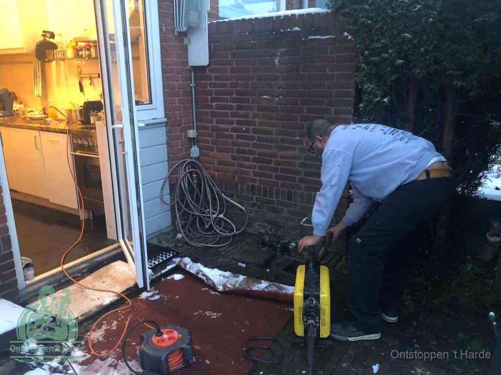 Een monteur werkt buiten bij een open riool-/grondput met een elektrische ontstoppingsmachine om een verborgen afvoerprobleem op te sporen in t Harde