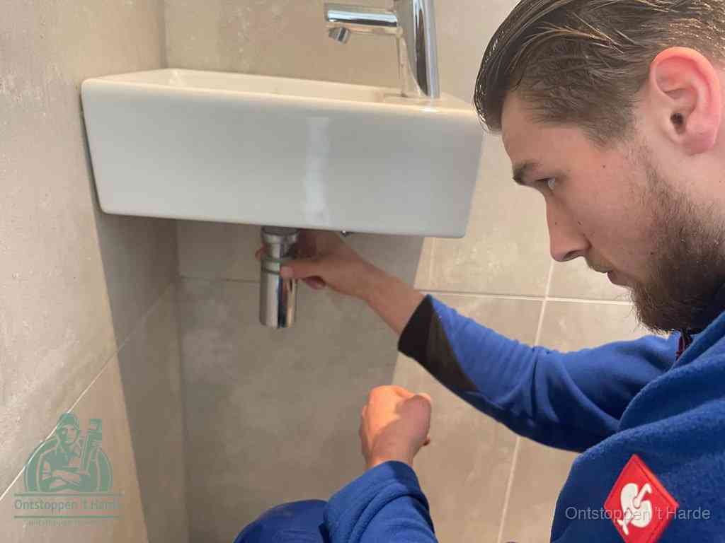 Een monteur draait met de hand de zwanenhalssifon onder een fonteinwasbak in de toiletruimte los om de bron van rioollucht in t Harde te vinden