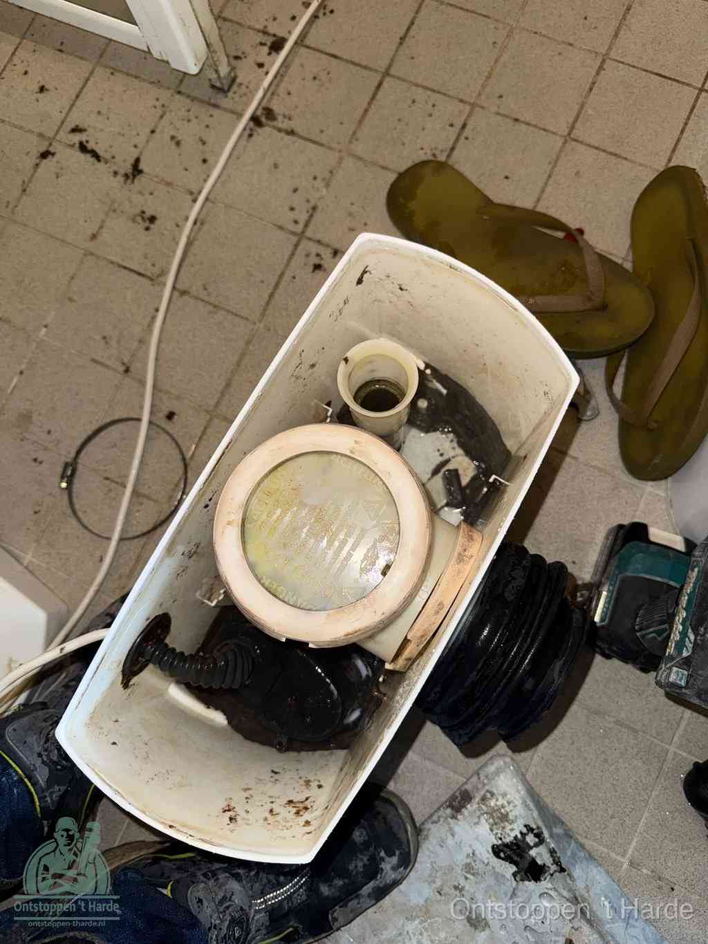 Een gedemonteerde toiletpot op de vloer met ontstoppingsveer en gereedschap ernaast, het verwijderen van een vastzittend voorwerp uit de wc in t Harde