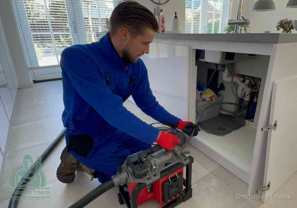 Een monteur voert een professionele elektrische ontstoppingsmachine in de afvoer onder de spoelbak tijdens het gootsteen ontstoppen in t Harde